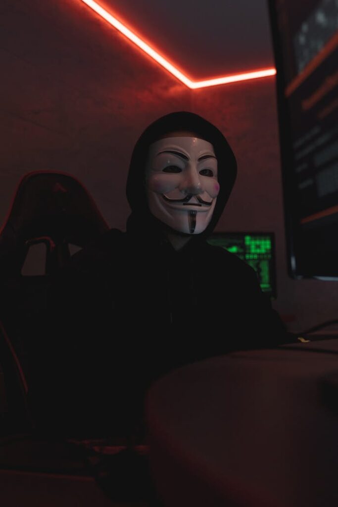 Mysterious figure wearing a Guy Fawkes mask, illuminated by computer screens in a dark room.